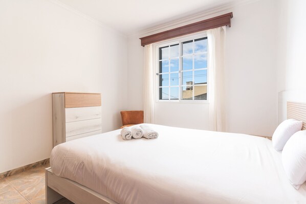 Ein ordentlich bezogenes Bett mit weißen Leinen und zwei aufgerollten Handtüchern, ein Holzkopfbrett, ein Fenster mit Blick auf Gebäude und eine weiße Wand.