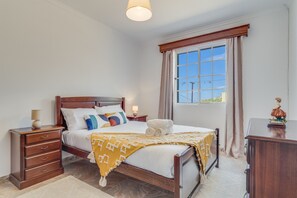Ein Schlafzimmer mit einem hölzernen Bett, einem Nachttisch mit Lampe, einer Kommode und einem Fenster mit Vorhängen.
