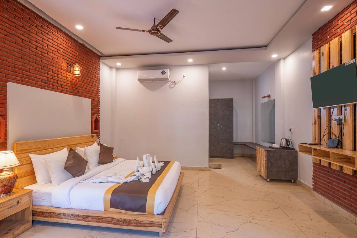 A hotel room with a brick wall, a bed with white bedding, a wooden nightstand, a lamp, a ceiling fan, an air conditioning unit, a TV, and a wooden shelf.