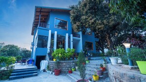 A modern two-story building with a blue facade, large windows, and a balcony. There are steps leading up to the entrance, surrounded by potted plants. - Destin Ekaant Resort by 3Tree Group (Ramnagar)