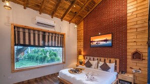 A bedroom with a wooden ceiling, a large window with a view of greenery, a bed with pillows and towels, a nightstand with a lamp, and a painting on the wall. - Destin Ekaant Resort by 3Tree Group (Ramnagar)
