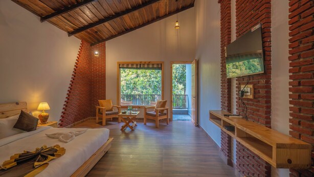 A bedroom with a bed, a desk, two chairs, a TV, and a balcony with a view of trees. - Destin Ekaant Resort by 3Tree Group (Ramnagar)