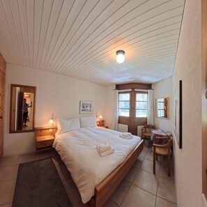 Ett sovrum med en säng i trä, sänglampor, en spegel, en inramad tavla och ett fönster med gardiner.