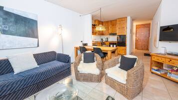 Een moderne woonkamer met een bank, twee fauteuils, een glazen salontafel, en een eethoek met een keuken op de achtergrond.
