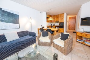 Ett modernt vardagsrum med en soffa, två fåtöljer, ett soffbord i glas och en matplats med ett kök synligt i bakgrunden.
