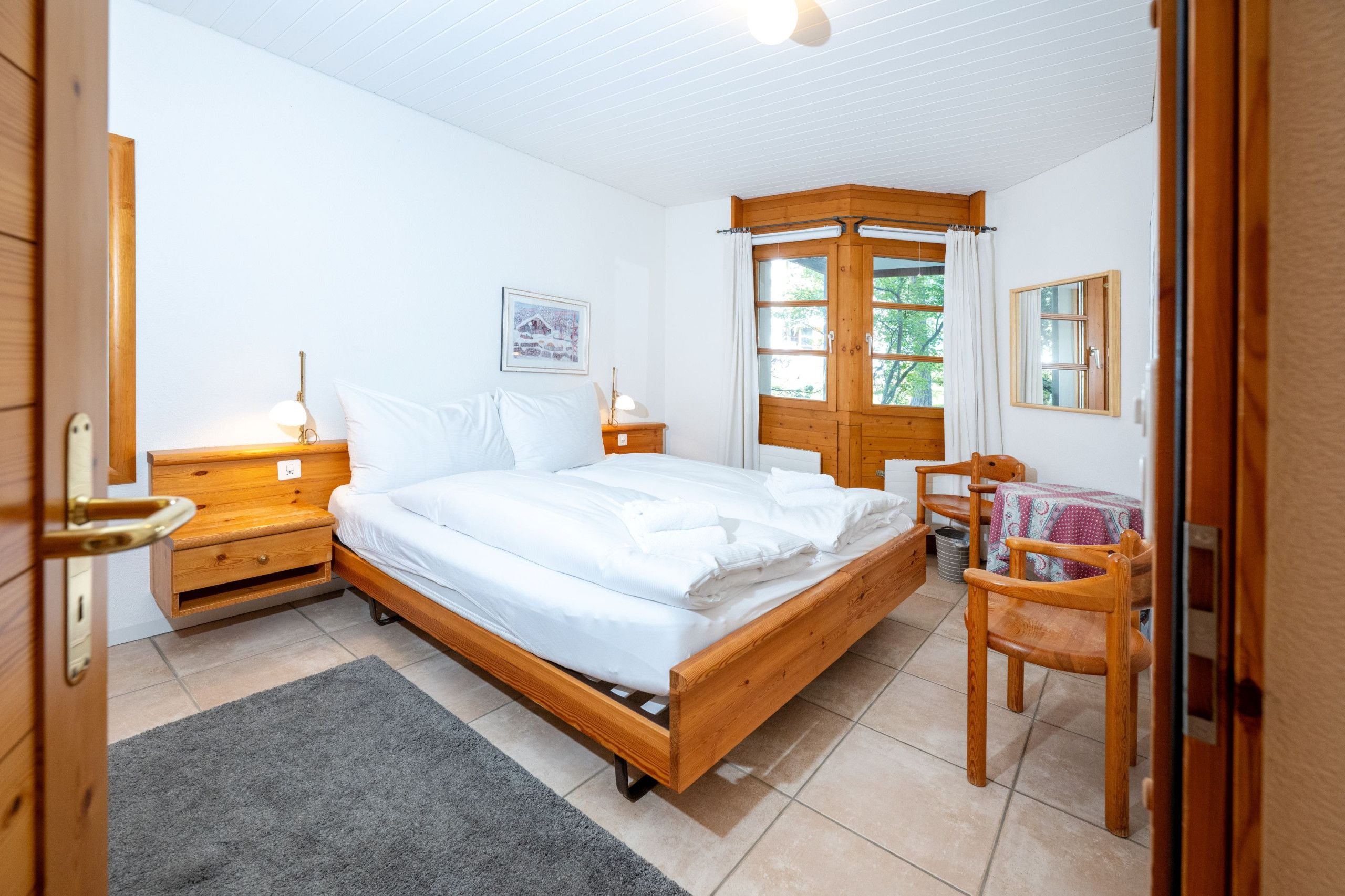 Une chambre à coucher avec un lit en bois, des tables de chevet, une chaise, une fenêtre avec des rideaux et un tableau encadré au mur.