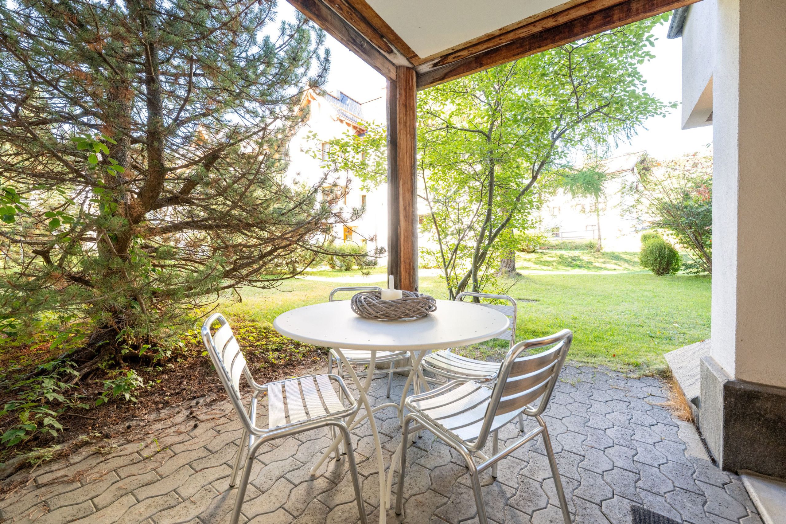 Eine Außenpatio mit einem runden Tisch und zwei Stühlen, umgeben von einem Garten und einem Gebäude.