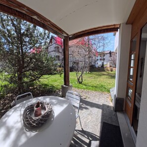 En täckt uteplats med ett bord och stolar, utsikt över en trädgård och en byggnad med ställningsmaterial.