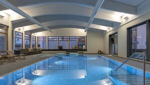 An indoor swimming pool with clear blue water, a lounge area with chairs, and large windows providing natural light. - Wellness apartment with pool and terrace (Lenzerheide/Lai)