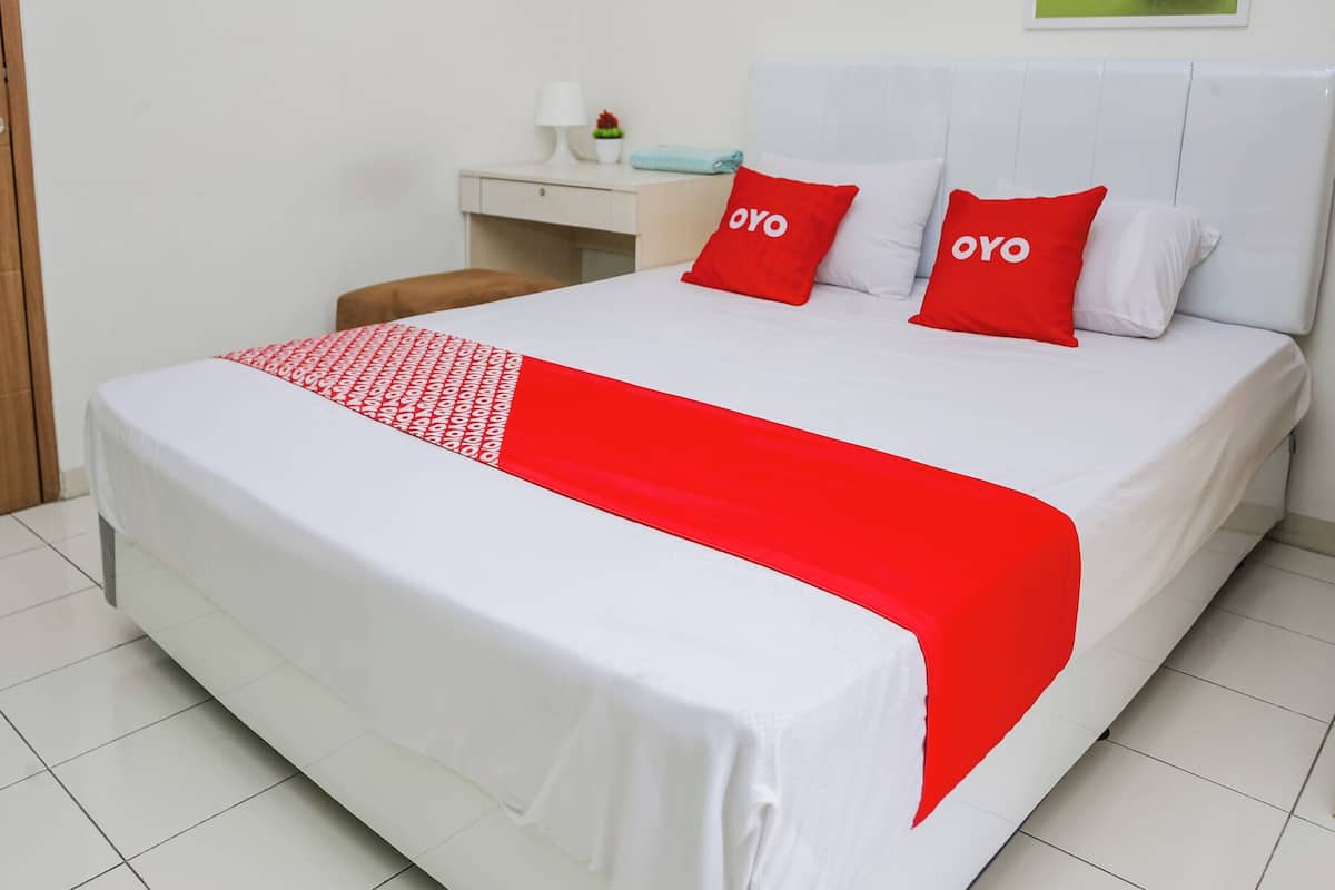 A neatly made bed with white and red bedding, a bedside table with a lamp and a plant, and a small stool.