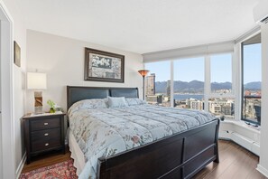 Quarto com uma cama grande, mesa de cabeceira, luminária, quadro e vista para a cidade através de janelas amplas.