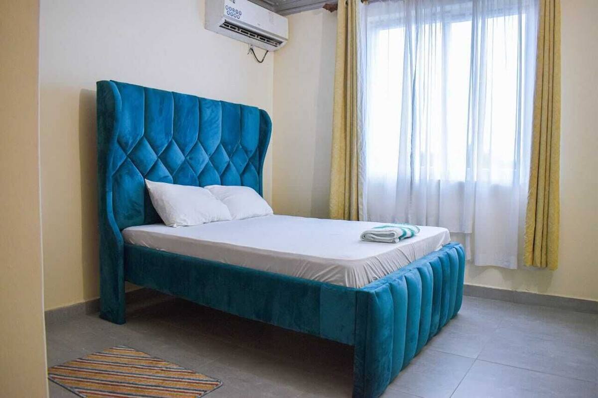 A bed with a blue headboard and white bedding in a room with a tiled floor and a window with curtains.