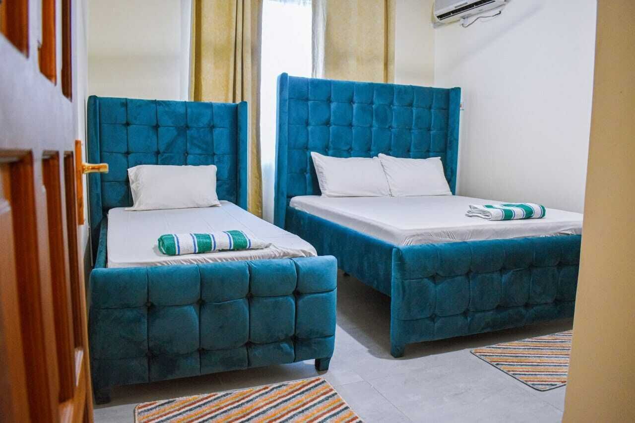 Zwei blaue Betten mit weißen Laken und gestreiften Handtüchern, ein Streifen Teppich und ein Fenster mit Vorhängen.