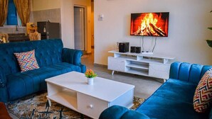 A living room with a blue sofa, a white coffee table, and a TV mounted on the wall displaying a fireplace. - Pavilion Suites Nyali (Mombasa)