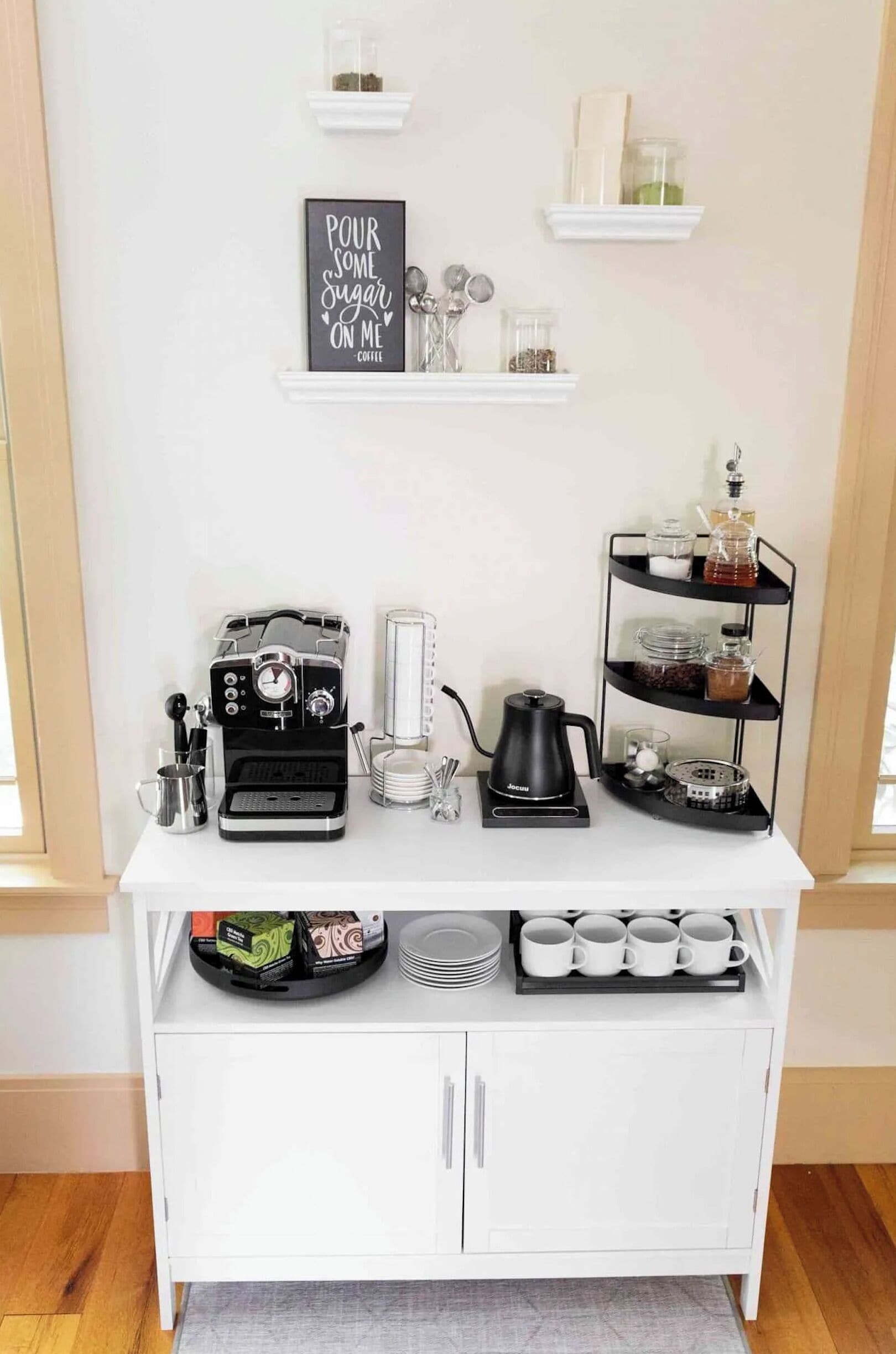 A white coffee station with a coffee maker, milk frother, and a shelf with coffee cups and containers.
