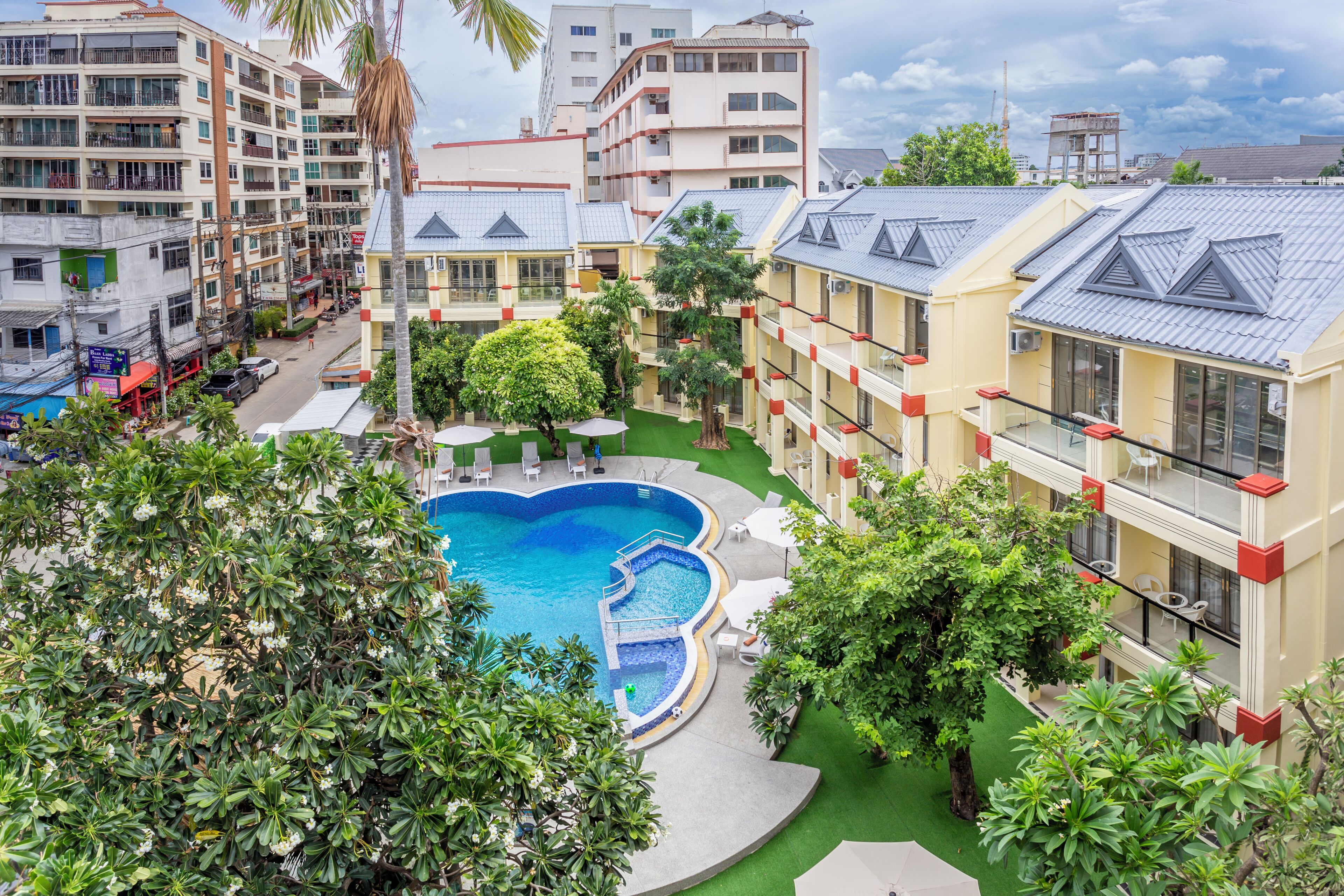 A hotel with a swimming pool surrounded by greenery, multiple floors, and a mix of residential and commercial buildings in the background.