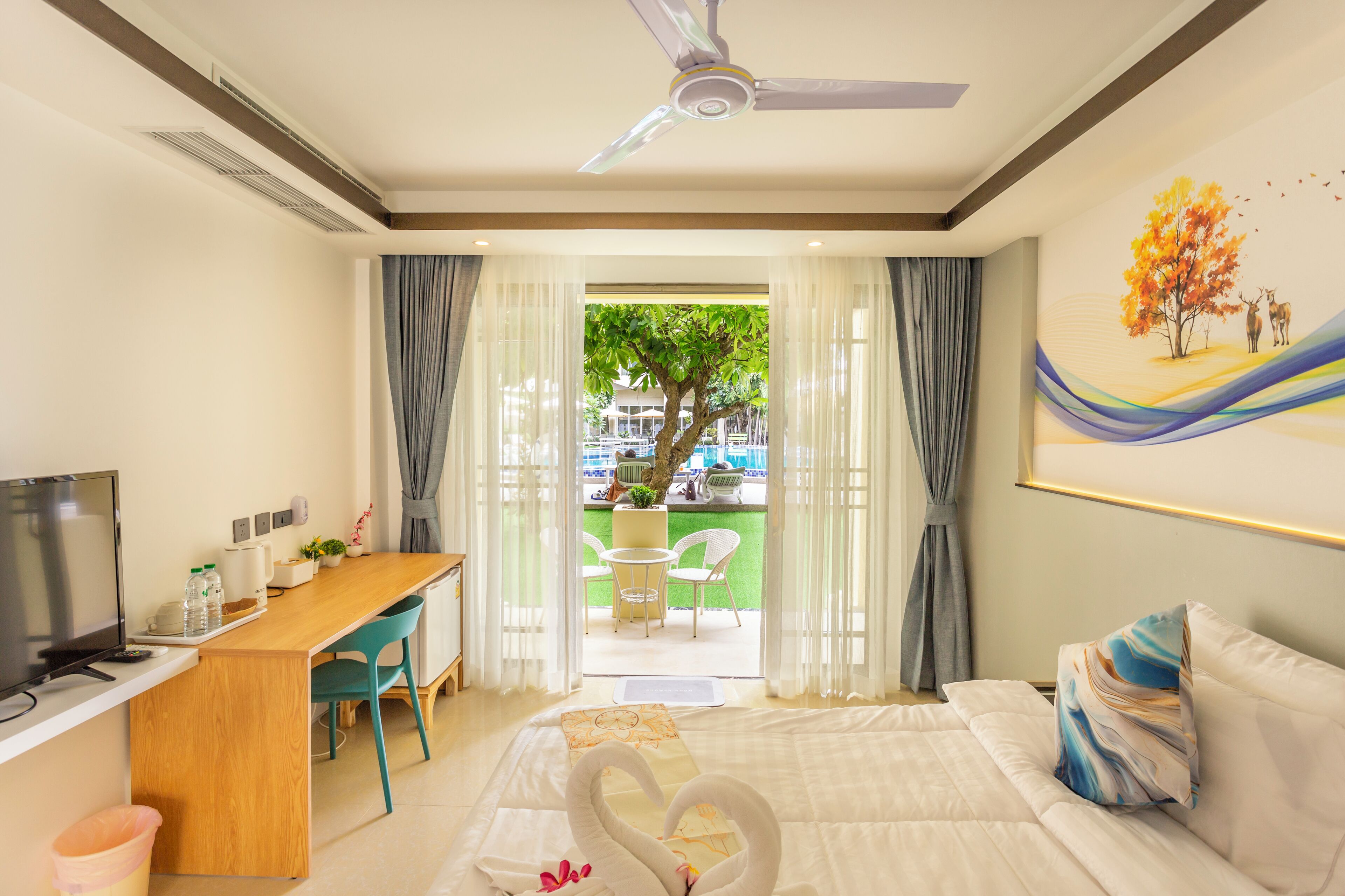 Habitación de hotel con cama, escritorio, televisor y vista a un espacio exterior con árbol y sillas.
