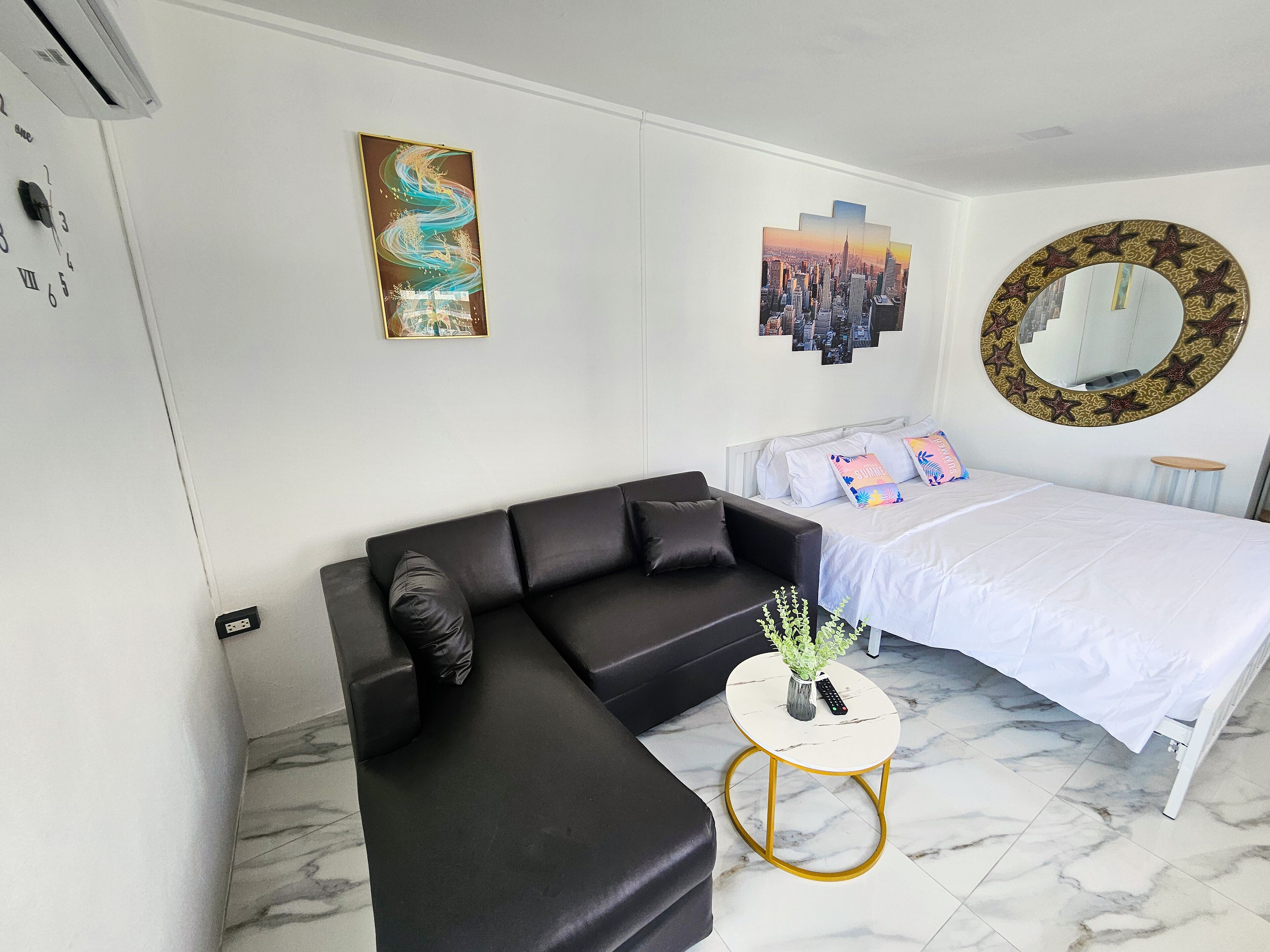 Une chambre moderne avec un canapé d’angle noir, un lit simple avec une couette blanche, un miroir rond orné de motifs en forme d’étoiles et une œuvre d’art représentant un paysage urbain accrochée au mur.