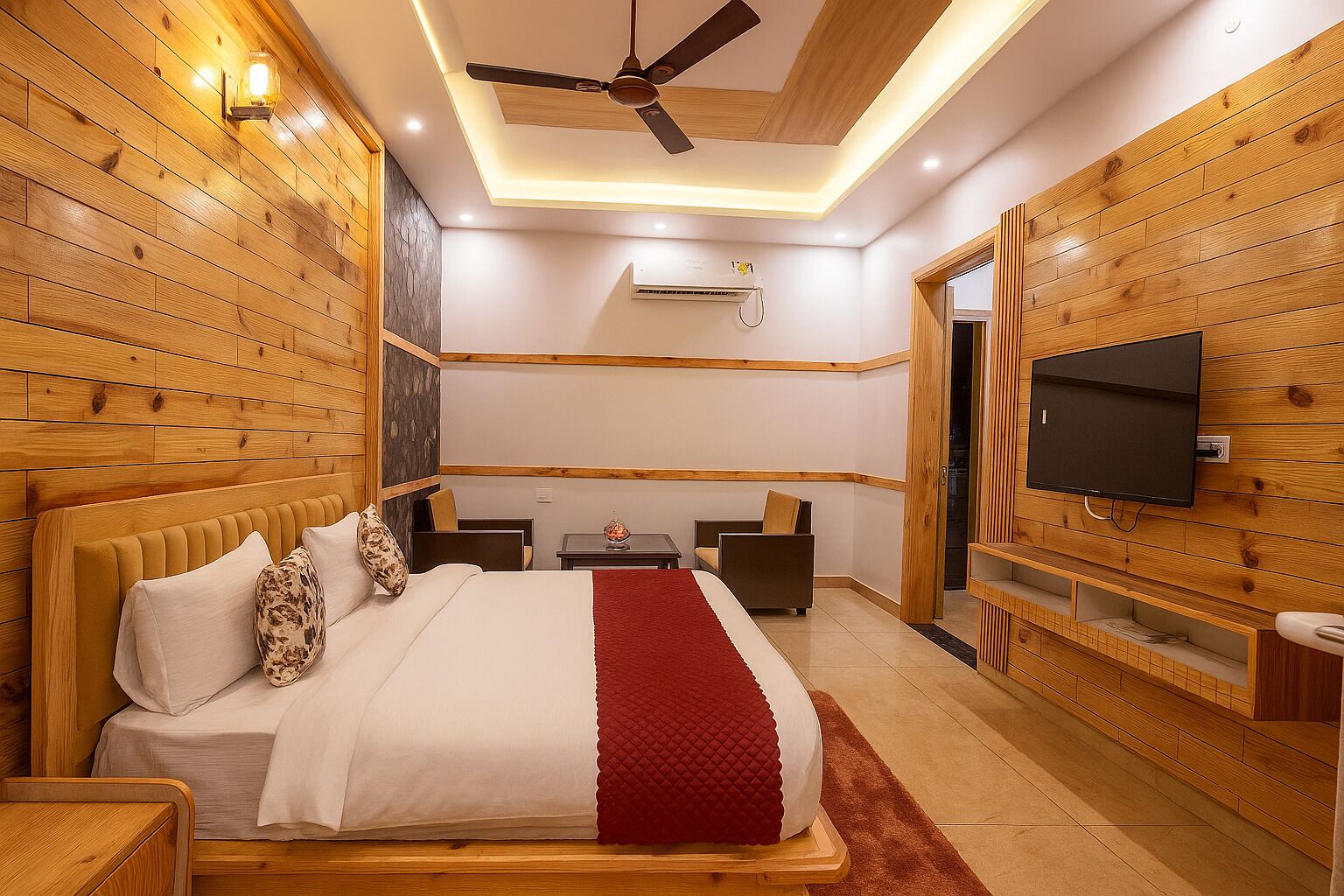 A modern bedroom with a wooden headboard, a flat-screen TV mounted on the wall, a ceiling fan, and a small table with a chair.