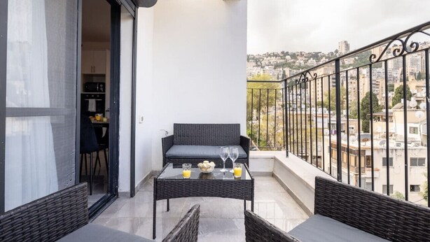 A balcony with wicker furniture, a glass table with drinks, and a city view. - MAMAD - Stella Maris views Vista apartments by Sea N' Rent (Haifa)