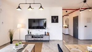 A modern living room with a flat-screen TV, a grey console table with decorative items, a dining area with a table set for two, and a kitchen in the background. - MAMAD - Stella Maris views Vista apartments by Sea N' Rent (Haifa)