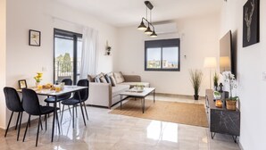 A modern living room with a dining area, a sofa, a coffee table, and a TV. There are plants and a lamp on a side table. - MAMAD - Stella Maris views Vista apartments by Sea N' Rent (Haifa)