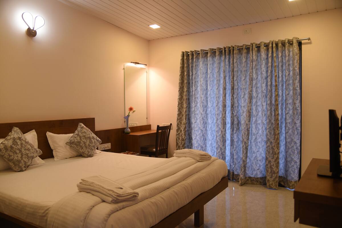 A neatly made bed with pillows, a bedside table with a lamp, a chair, and a large window with curtains.