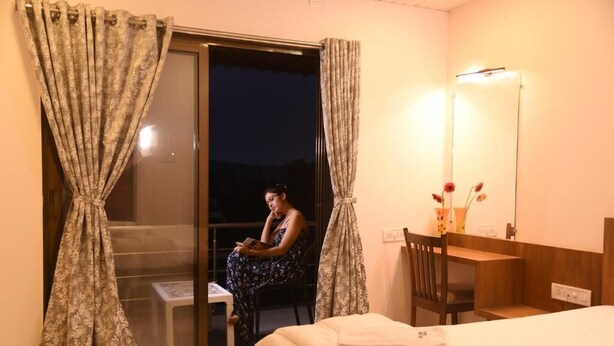 A hotel room with a bed, a desk, a chair, a table, and a sliding glass door leading to an outdoor area. - Stone Shelter (Mahabaleshwar)