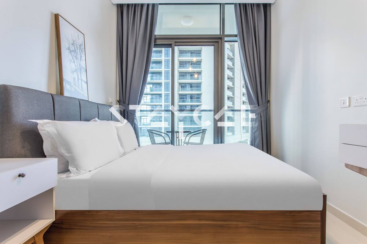 A modern hotel room with a large bed, a bedside table, a chair, and a view of a high-rise building through a window.