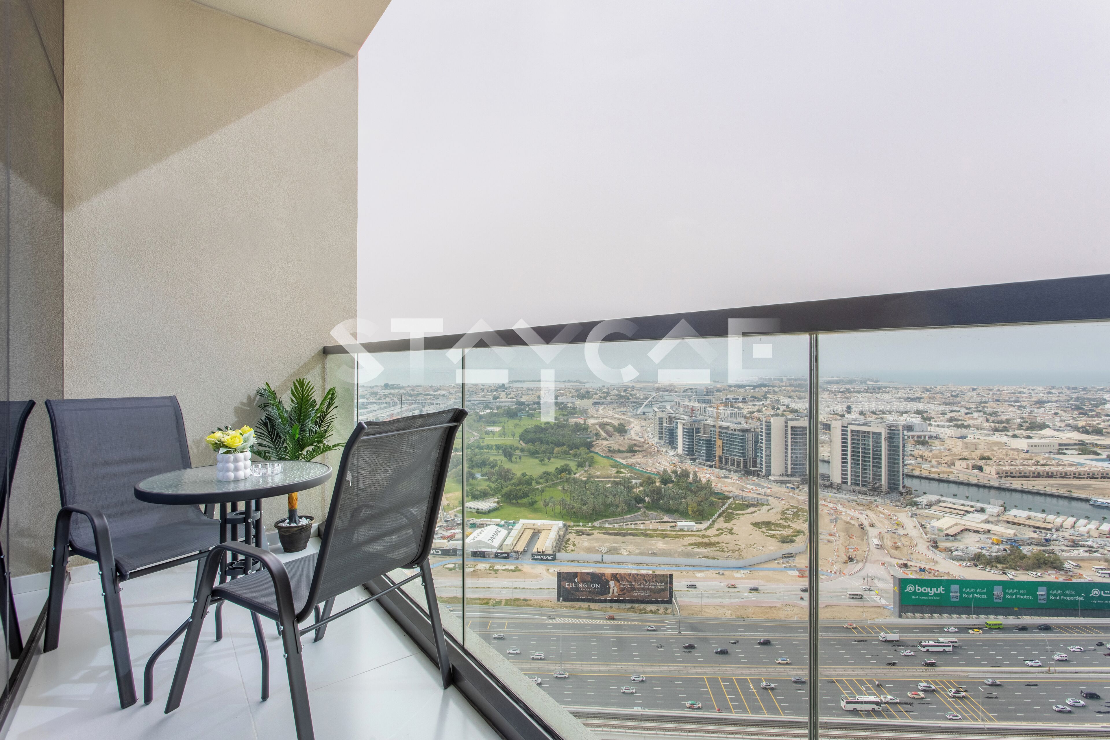 Un balcon avec une table et des chaises, un pot de fleurs et une vue sur un paysage urbain avec des immeubles et une autoroute.
