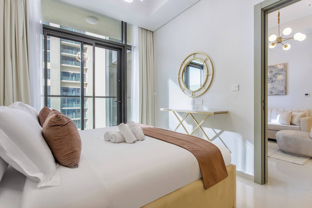 A modern hotel room with a large bed, a bedside table, a mirror, and a view of the city through large windows.