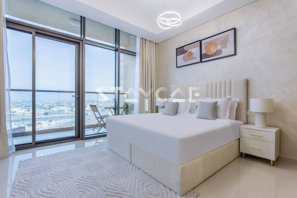 A modern bedroom with a large bed, a bedside table, a view through floor-to-ceiling windows, and a cityscape.