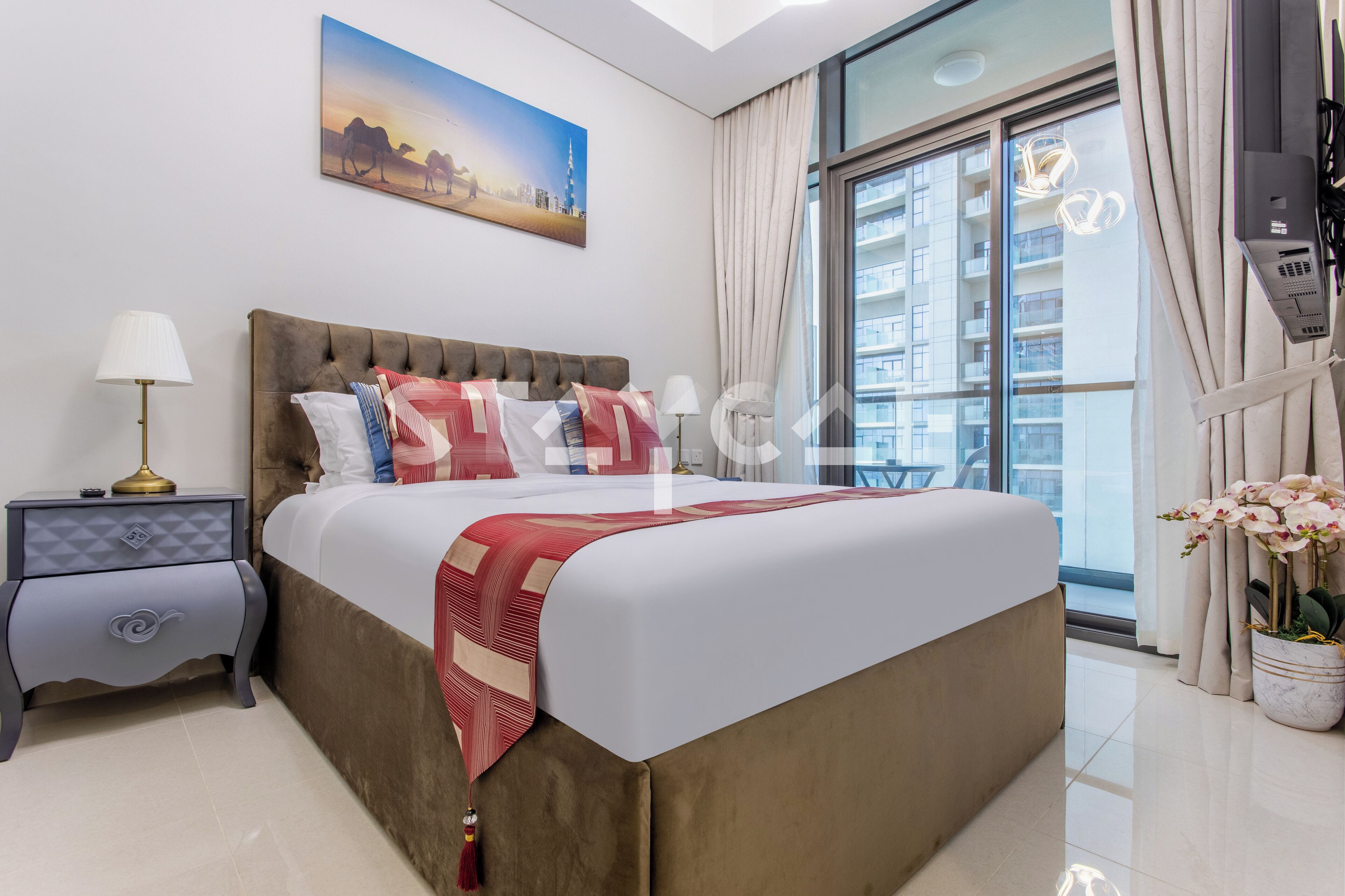 Une chambre d’hôtel moderne avec un grand lit, une table de chevet, une lampe, un tableau, une plante en pot et une vue sur un immeuble de grande hauteur.