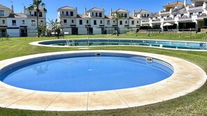 Una piscina con borde de azulejos, rodeada de césped y una hilera de edificios blancos con tejados rojos.