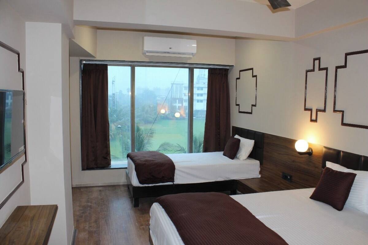 A hotel room with two beds, a large window with curtains, a flat-screen TV, and a wall-mounted light fixture.