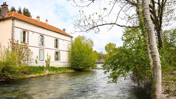Une rivière coule le long d’un bâtiment dont le toit est recouvert de tuiles rouges et qui possède des volets verts.