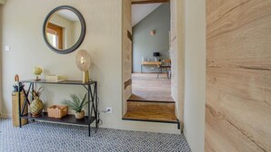 Un couloir doté d’un plancher en bois, une console noire ornée d’objets décoratifs, un miroir rond et un escalier menant à une pièce où se trouvent un bureau et une chaise.