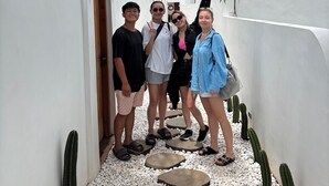 Un grupo de personas caminando por un sendero estrecho flanqueado por paredes blancas con barrotes de ventanas negros, piedras y cactus.