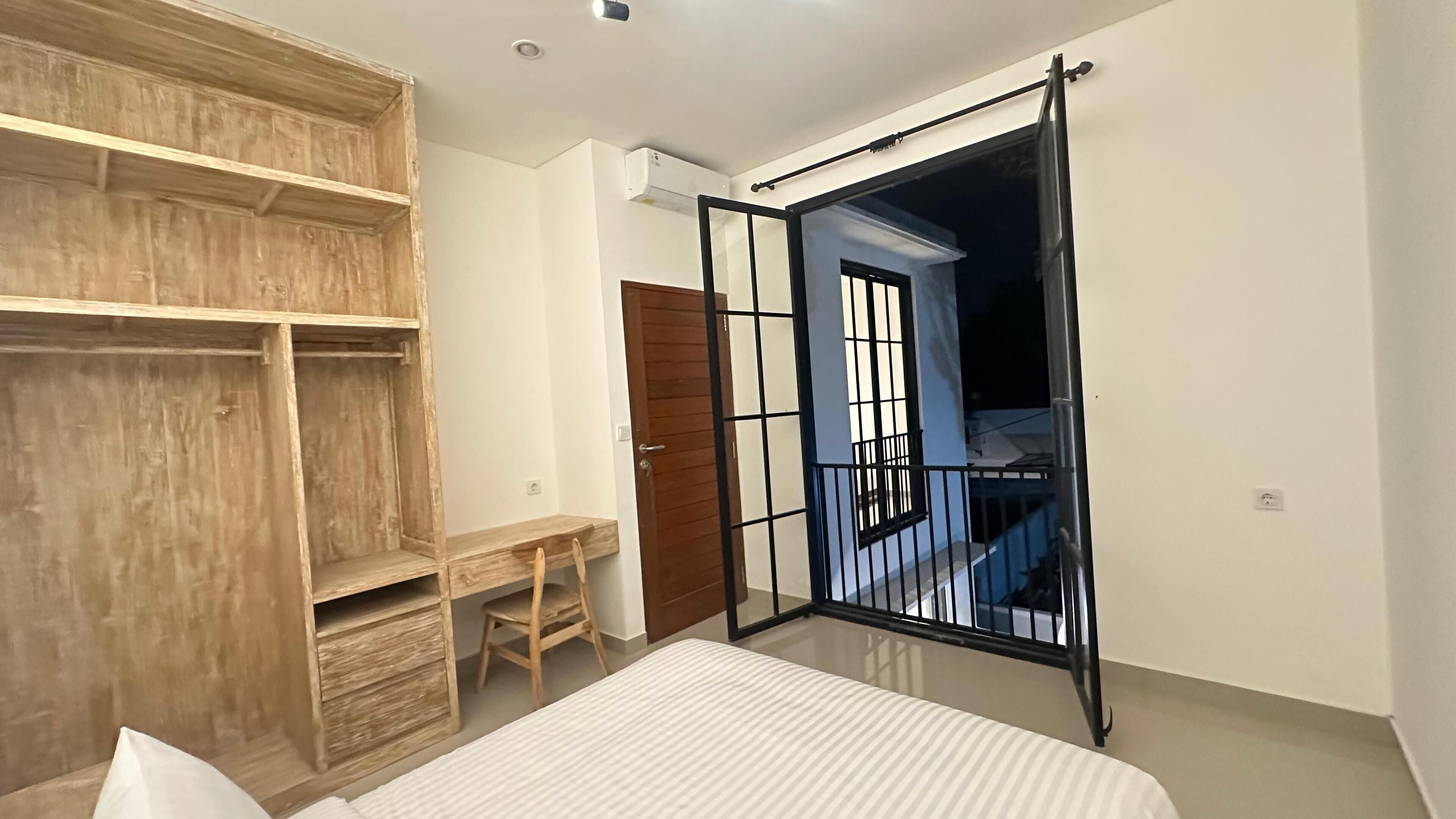 A bedroom with a bed, a wooden wardrobe, a desk, and a balcony with a black railing.