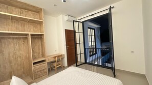 A bedroom with a bed, a wooden wardrobe, a desk, and a balcony with a black railing.