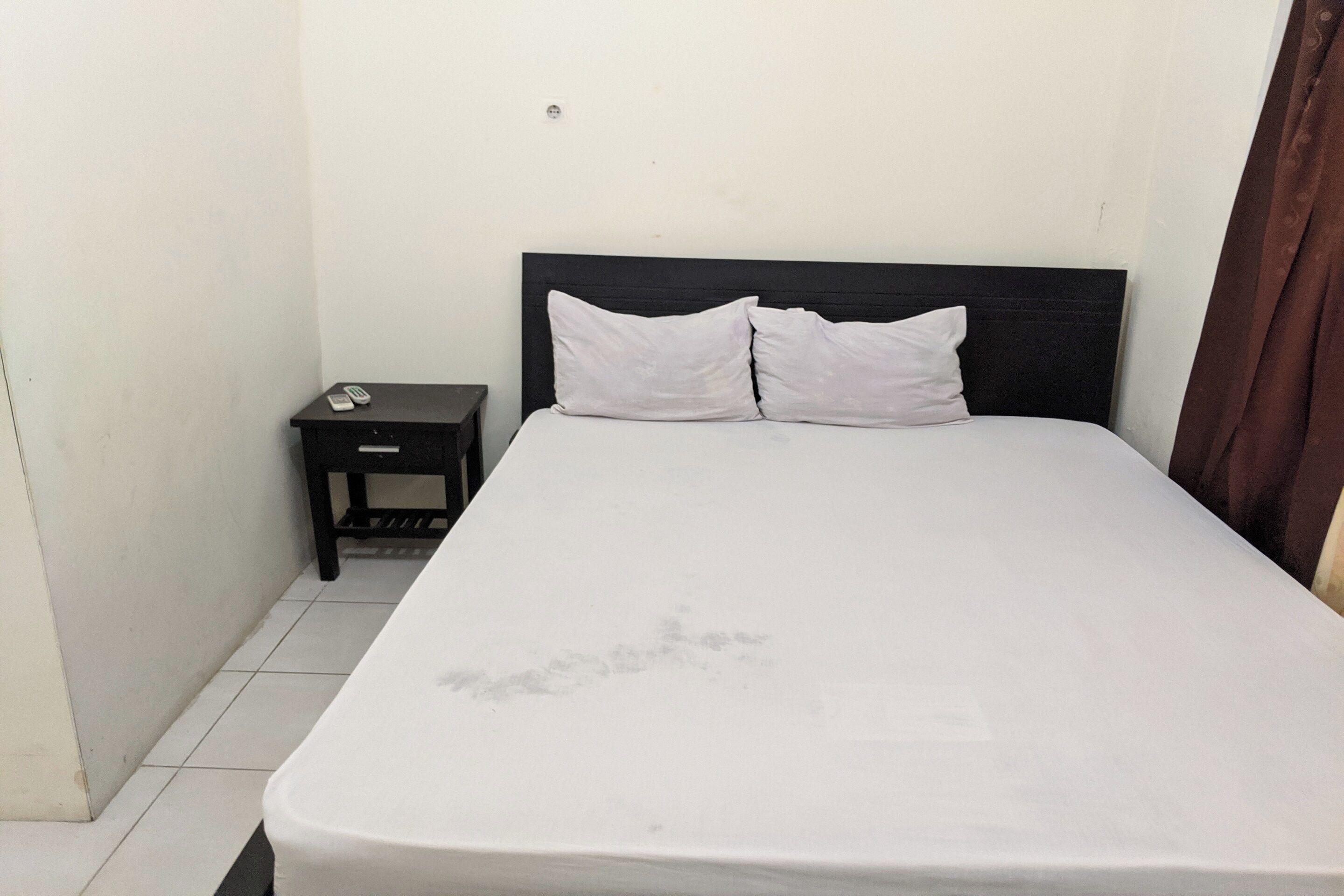 Ein Einzelbett mit dunklem Kopfteil und weißer Bettwäsche in einem kleinen, schlichten Zimmer mit weißen Wänden und Fliesenboden.