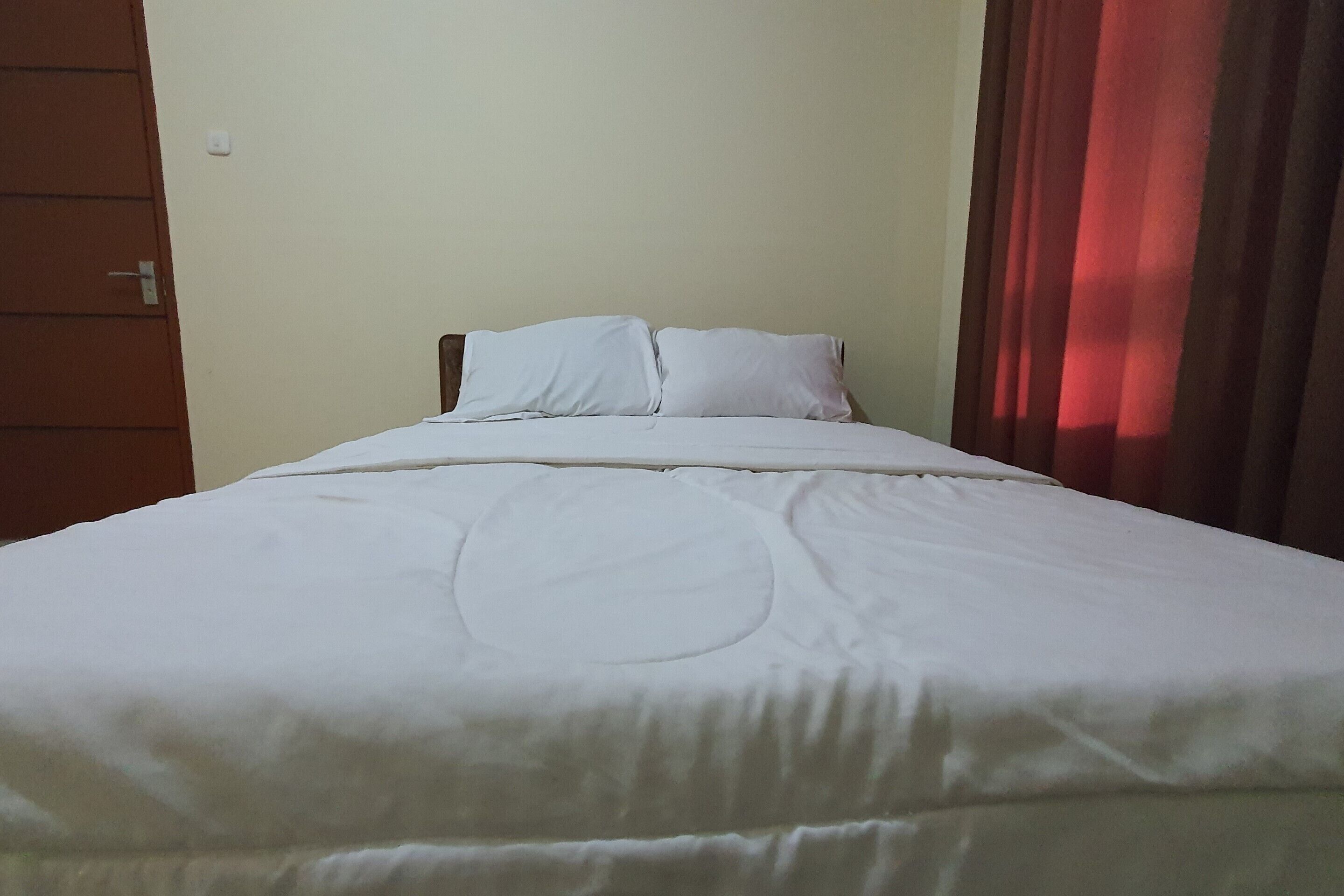 A neatly made bed with white linens and a circular design on the bedspread, a wooden headboard, and a red curtain partially drawn to the side.