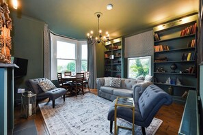 A living room with a sofa, armchairs, a coffee table, a chandelier, a fireplace, a bookshelf, and a piano. - Fairview Apt B - 2BR Gem in West Bridgford Centre (West Bridgford Nottingham)