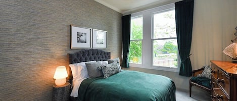 A bedroom with a large bed, a bedside lamp, a dresser, and a window with curtains.