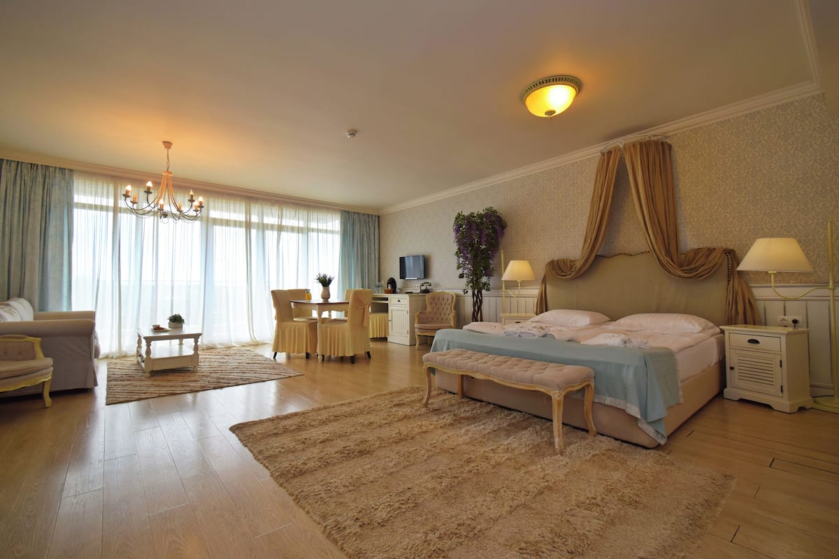 A spacious hotel room with a large bed, a sitting area with a sofa and armchairs, a dining table, and a television.