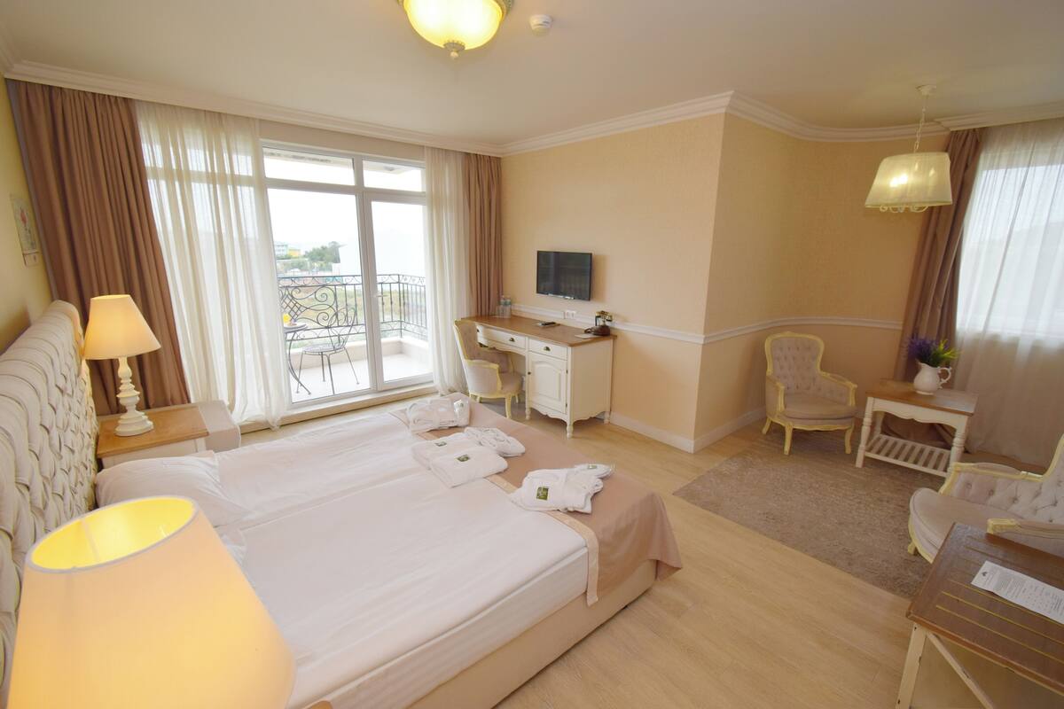 A hotel room with a large bed, a desk, a chair, and a balcony with a railing.