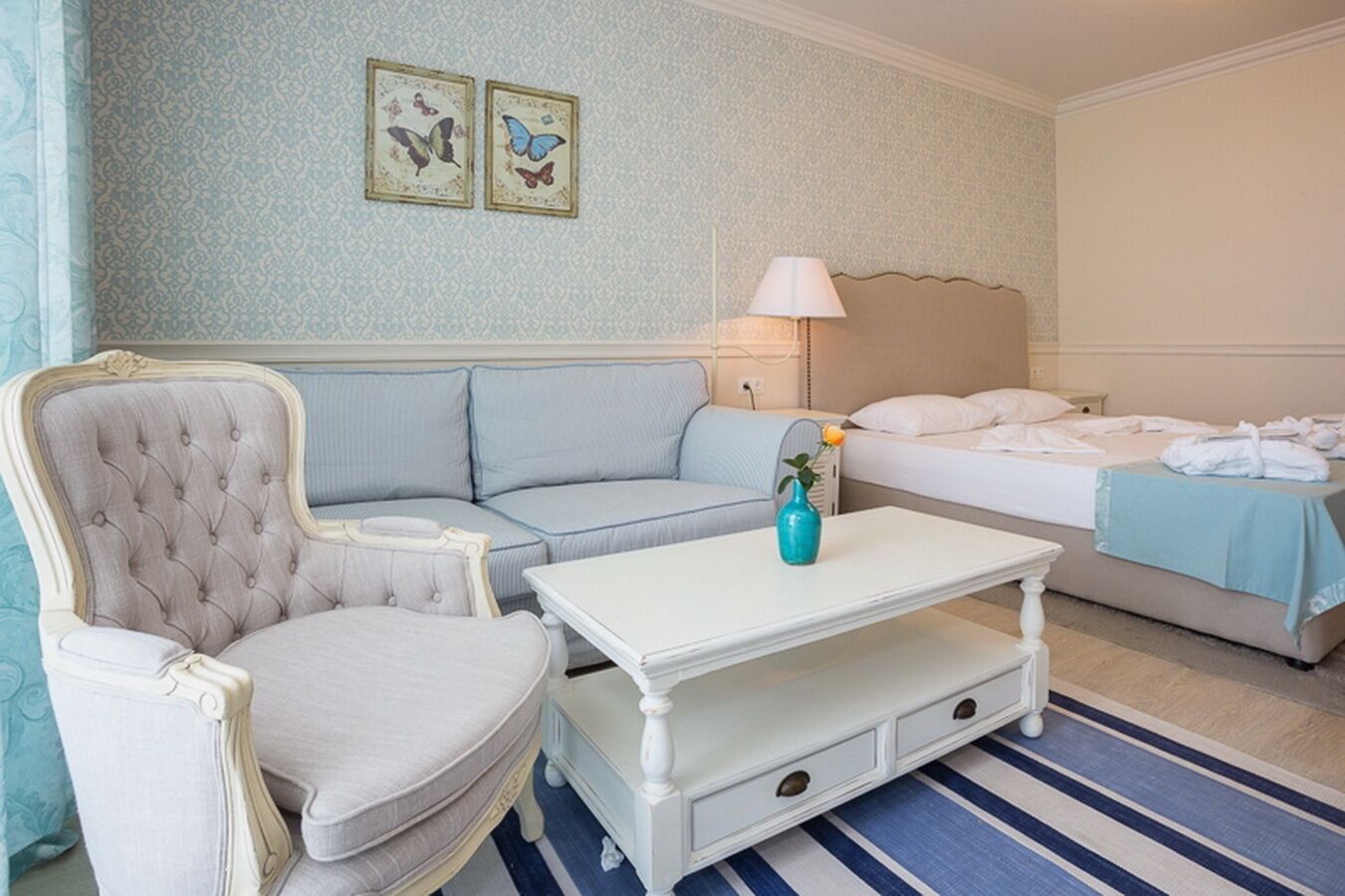 Una habitación de hotel con un sillón capitonado, un sofá, una cama, una mesita de noche, una lámpara y un florero con una flor.