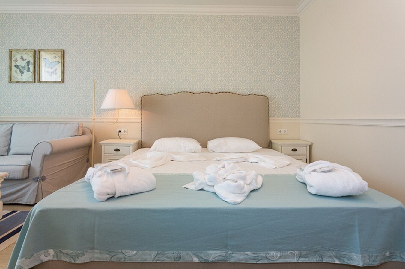 A hotel room with a bed, two bedside tables, a nightstand, a lamp, a sofa, and two framed pictures on the wall.