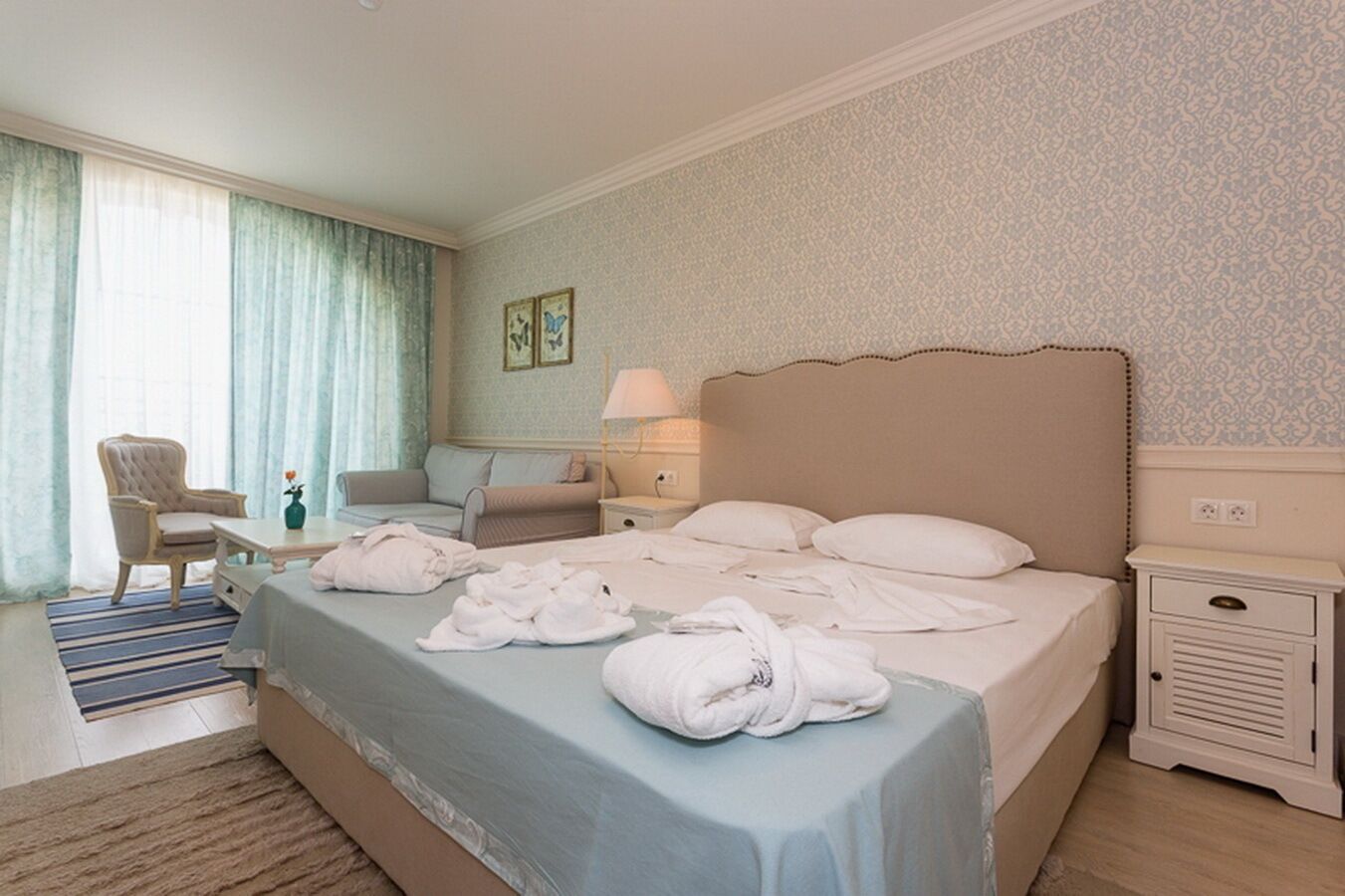 A hotel room with a large bed, a sitting area with a sofa and armchair, a small table with a lamp, and a window with curtains.