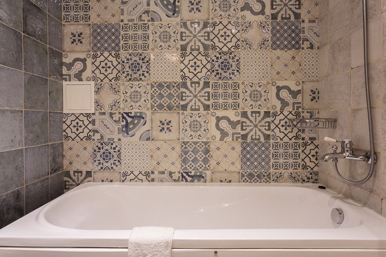 A bathroom with a white bathtub, tiled walls with various patterns, and a shower area with a handheld showerhead.
